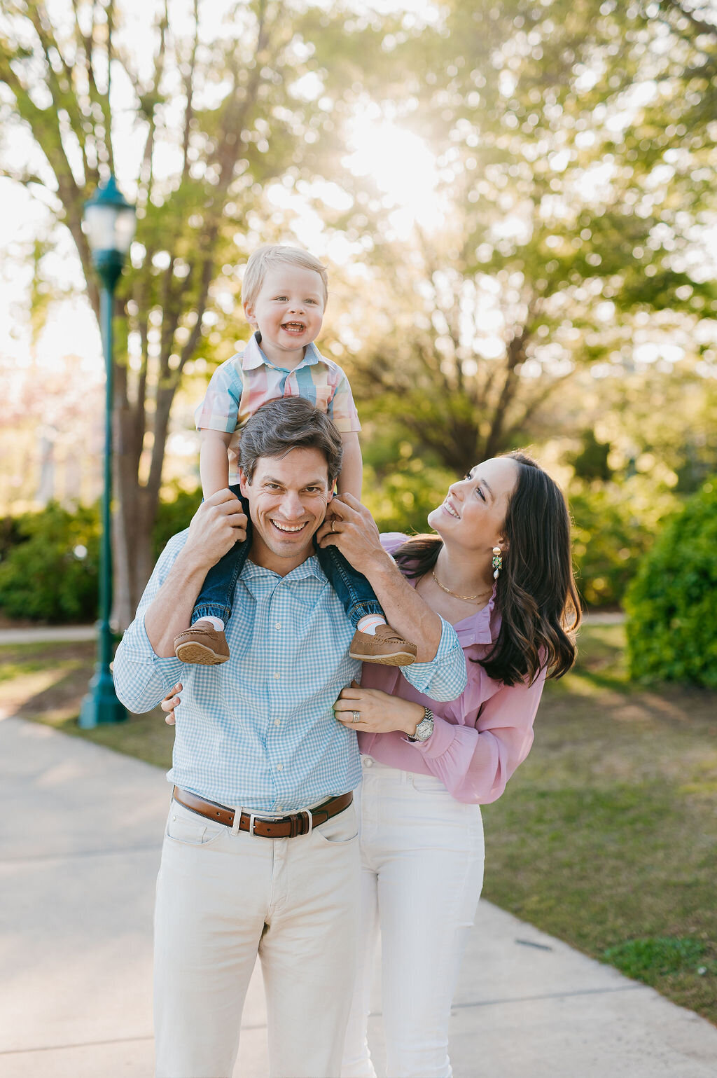 Parents laughing on a sunlit park path with their toddler son riding on his father's shoulders